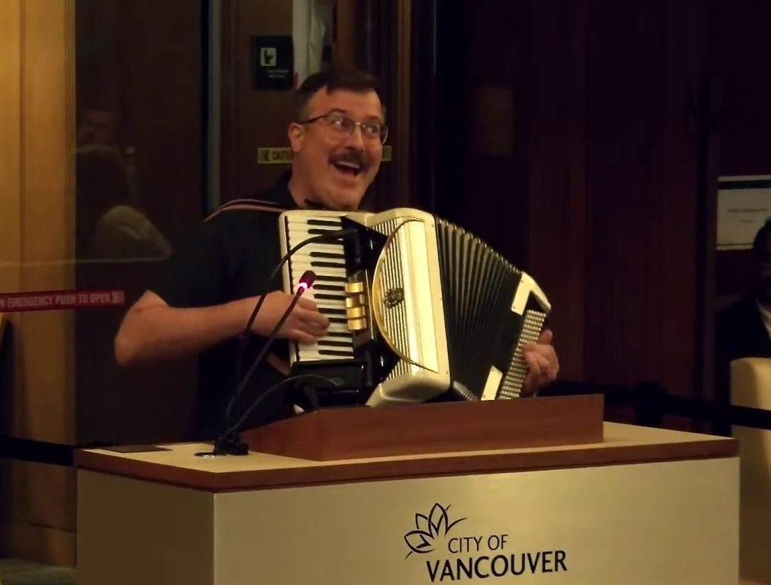 Photo of Peter Waldkirch playing an accordion at the speaker's podium at Vancouver City Hall.